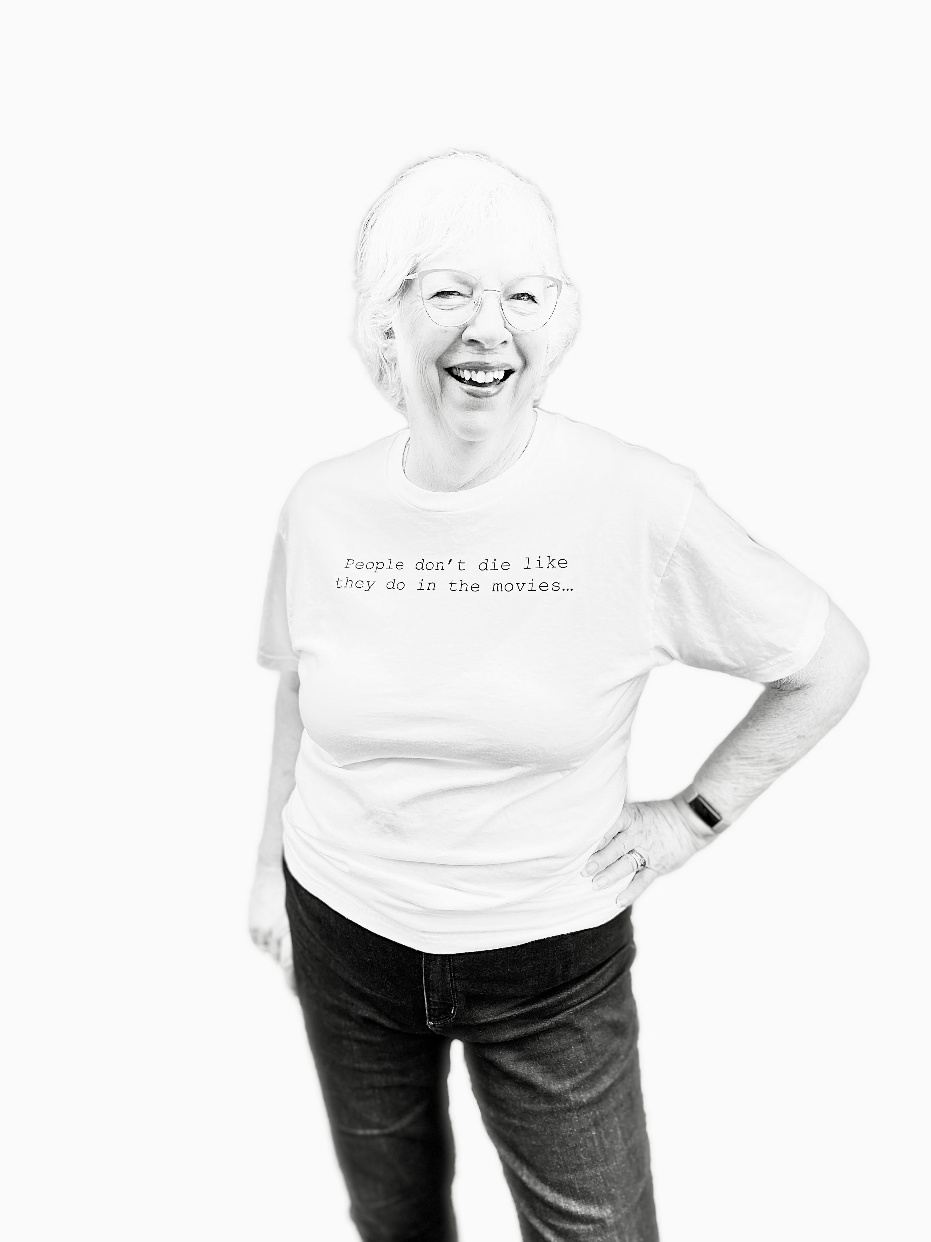 Barbara Karnes, RN wearing a white t-shirt with text that says her quote, People Don't Die Like They Do in the Movies, standing against a white background.