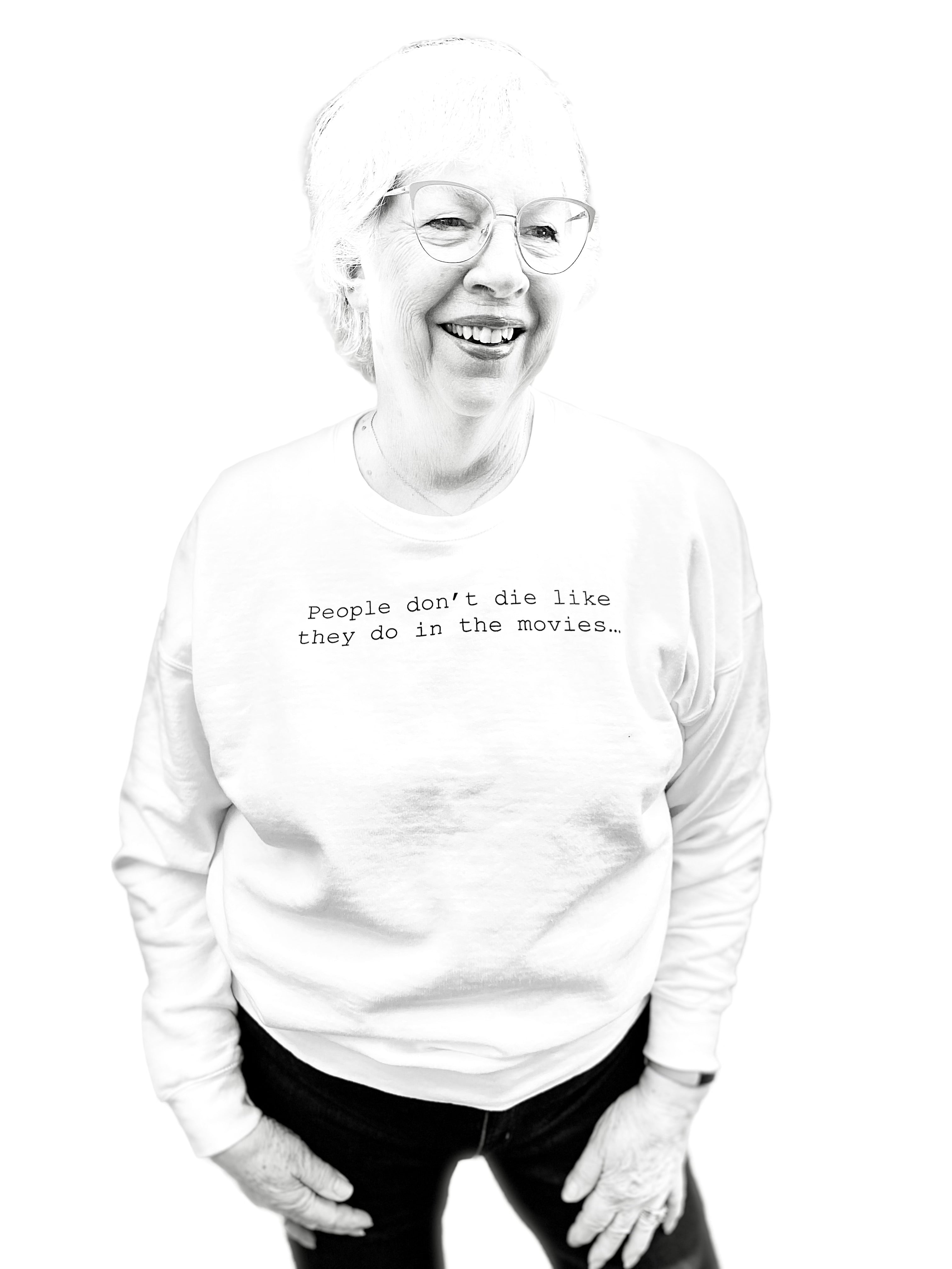 Barbara Karnes, RN wearing a white sweat-shirt with text that says her quote, People Don't Die Like They Do in the Movies, standing against a white background