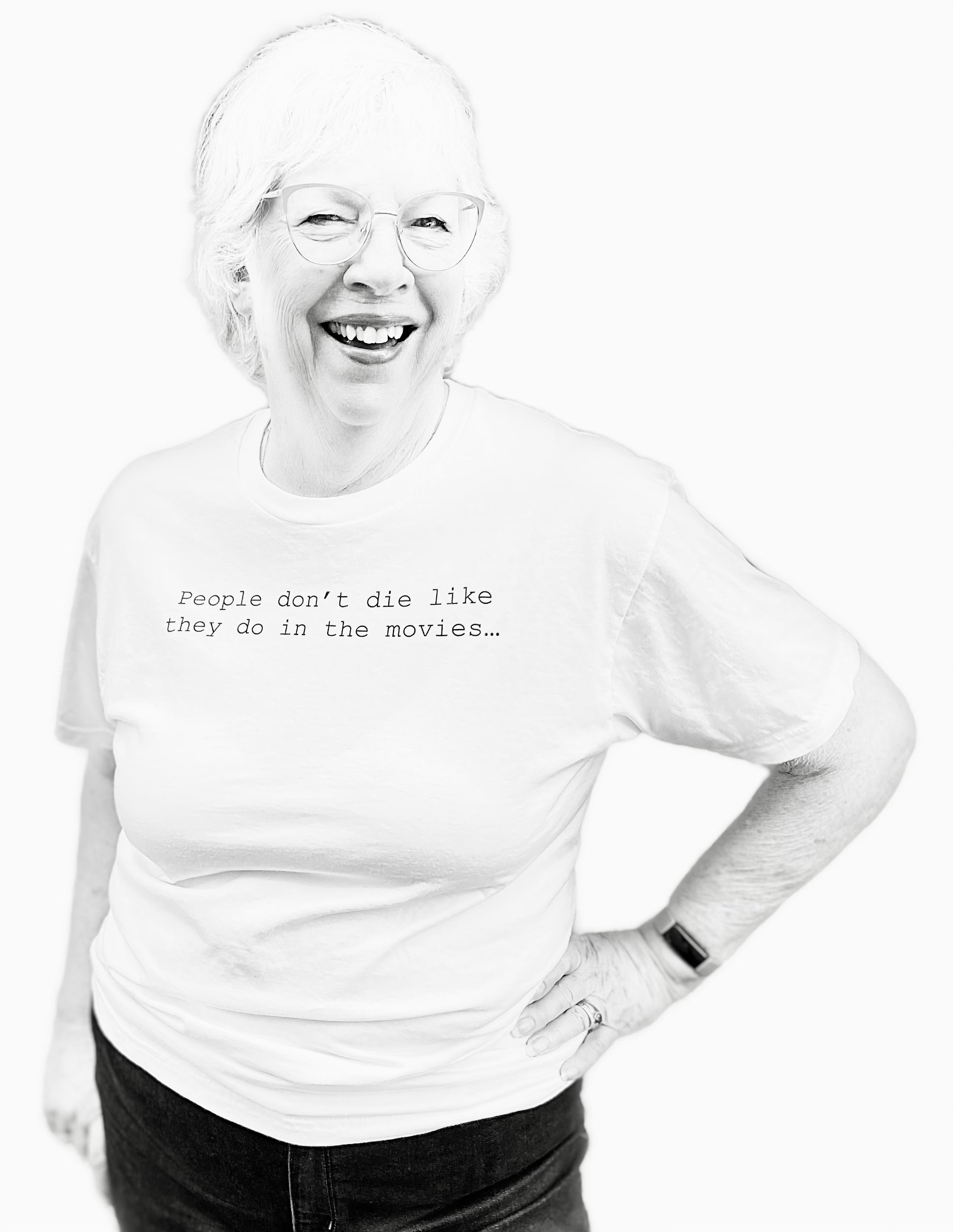 Barbara Karnes, RN wearing a white t-shirt with text that says her quote, People Don't Die Like They Do in the Movies, standing against a white background. Cropped.